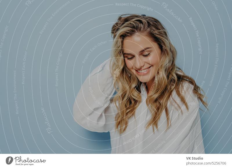 Romantic young blonde woman looking down with tender smile pleased caucasian romantic shy broadly smiling white casual shirt face beautiful wavy hair female