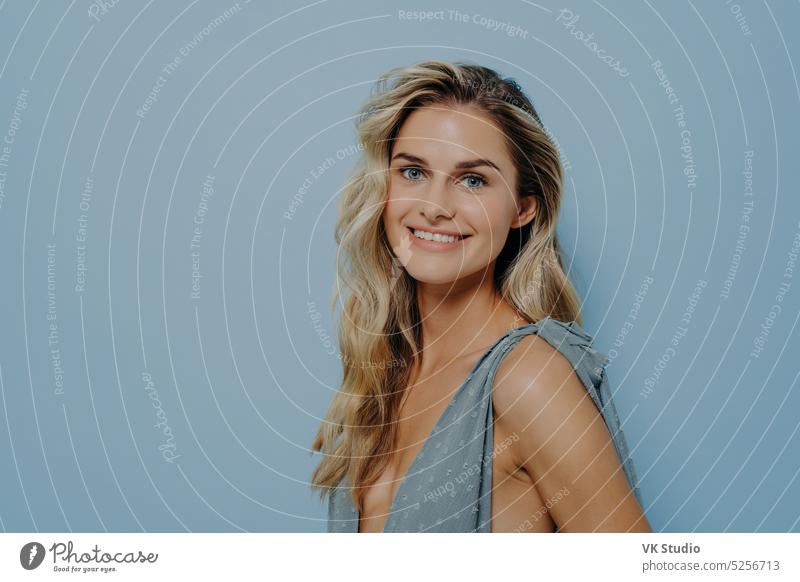 Cheerful smiling blonde girl in blue dress posing in studio cheerful woman beauty fashion standing sideways toothy smile looking calm relaxed time face isolated