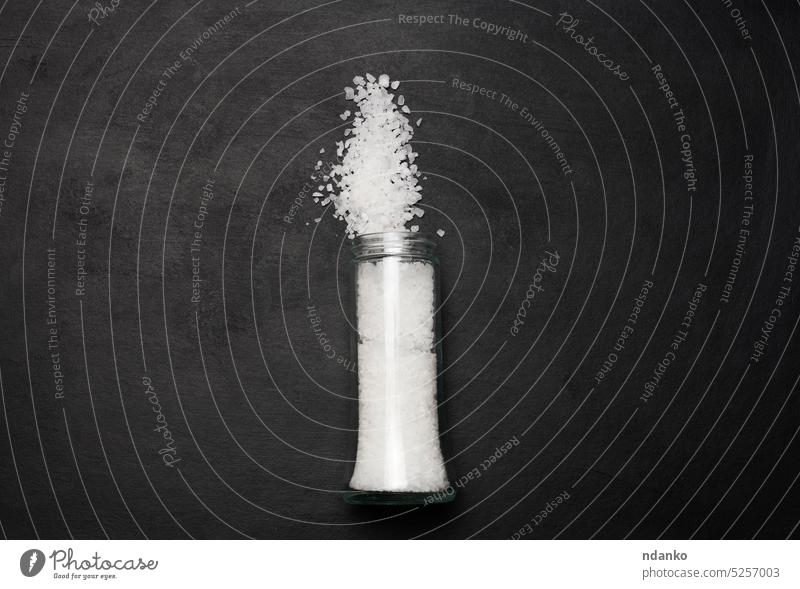 Large crystals of salt in a glass transparent bottle are scattered on a black background, top view white ingredient kitchen container food table spice mineral