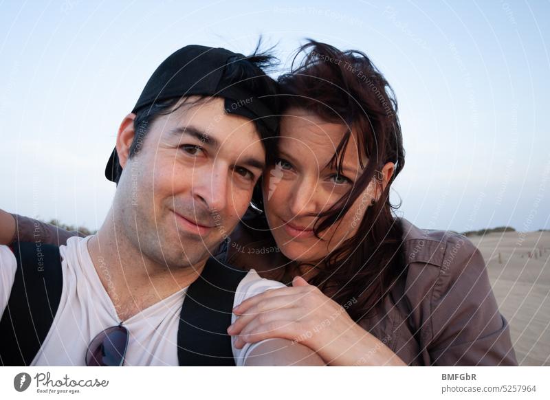Couple-er with baseball cap on backwards, they with windswept hair pose and cuddle for photo in desert. Woman Man couple Desert Cuddling windzersaust middle age