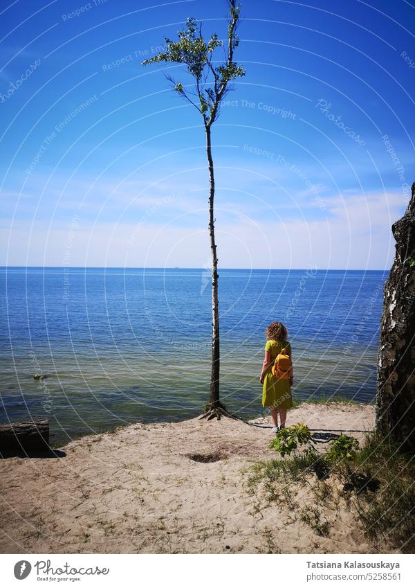 A curly-haired woman in a green dress with a yellow backpack stands on the shore of the Baltic Sea in Palanga and looks into the distance towards the horizon from a steep bank. A lone tree grows on the sand nearby.