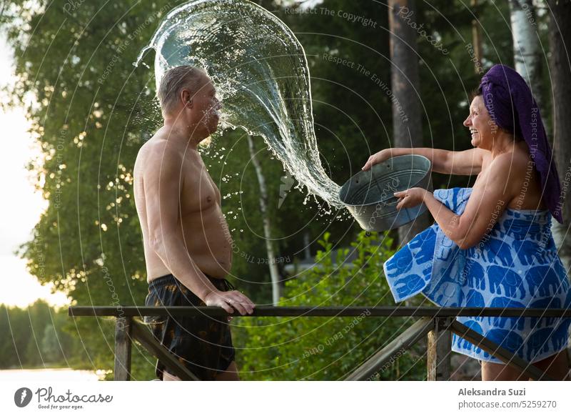 Aged couple having fun after Finnish sauna on wooden cottage pier in a lake. Mature woman pouring cold water from basin over her partner. Typical Finnish summer.