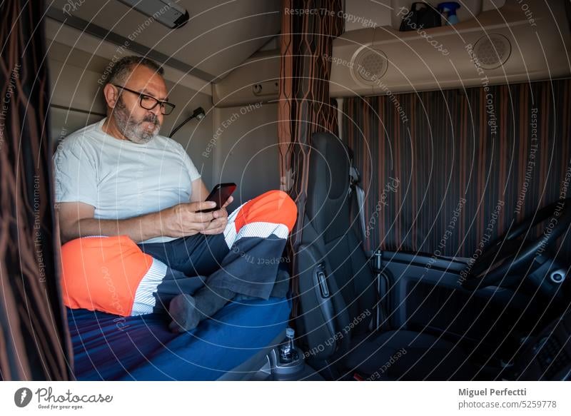Senior trucker sitting in the bed of the truck with the curtains drawn checking the mobile in his off-time. truck driver cabin profession man interior