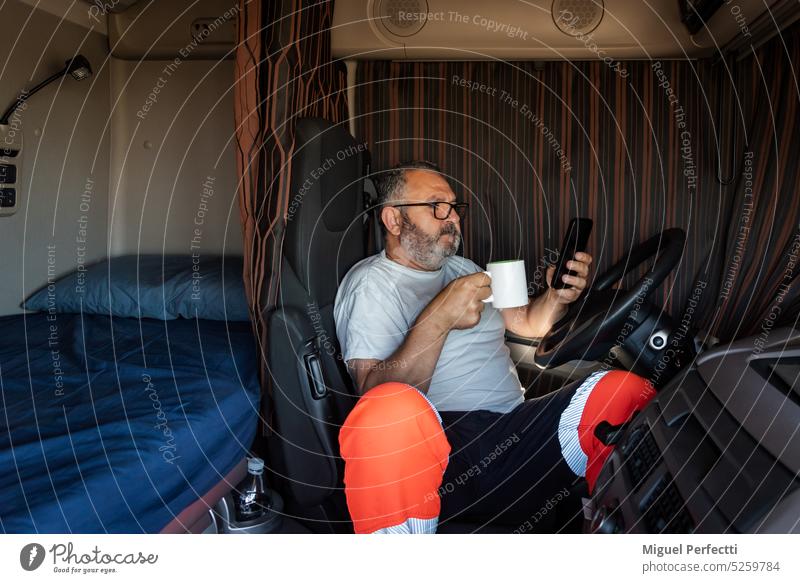 Senior trucker in the cab of the truck with the curtains drawn drinking morning coffee and looking at the phone. truck driver cabin older profession man inside