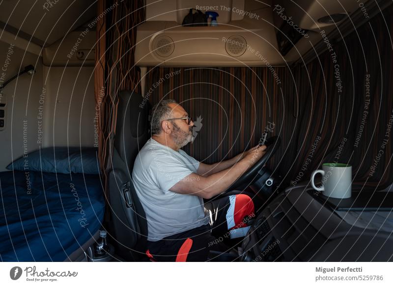A trucker in the cab, scrolling on his phone during his off-time, with the truck blinds down. truck driver cabin older profession man inside sitting coffee cup