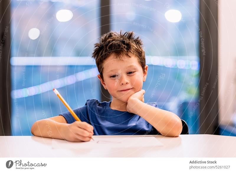 Glad boy sitting at table and drawing smile home window positive creative hobby evening child lean on hand happy cheerful glad childhood kid pencil sketch cute