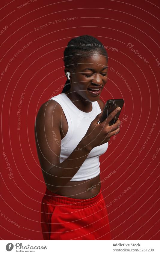Cheerful ethnic woman in activewear with smartphone near red wall cheerful happy listen music earphones using headphones smile model african american gadget