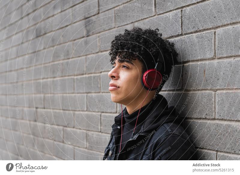 Stylish Latin American migrant listening to music on headphones man student brick wall building playlist street guy hispanic ethnic latin american colombia