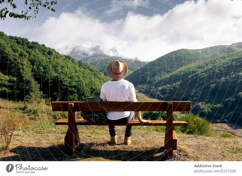 Male tourist resting on bench in mountains man traveler backpack admire destination countryside tourism young daytime holiday harmony straw hat casual adventure