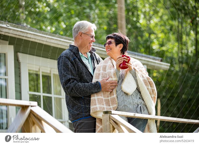 Mature couple standing on porch and drinking hot tea from mug. They are smiling and laughing, wrapped in a blanket. Happy senior couple embracing each other on the wooden terrace of the house.