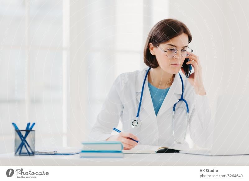 Heathcare personnel, medicine concept. Serious brunette female doctor focused at modern laptop computer, rewrites necessary information, talks on mobile phone, calls someone, has serious look