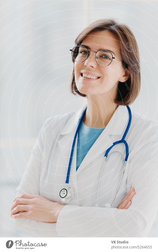 Close up portrait of short haired female general practitioner stands with smile and arms crossed, uses stethoscope, enjoys work, poses against white background. People, mediccare and treatment concept