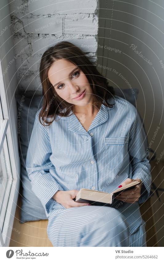 Relaxed young of beautiful in casual clothes, sits at window near brick wall, back to soft pillow, reads interesting detective story, looks directly at camera, wears casual costume. People and reading