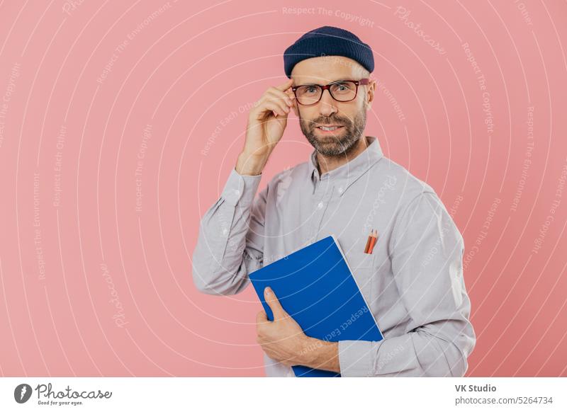 Stylish distant worker wears transparent glasses, stylish hat, shirt, holds blue textbook, uses pencils for writing information, poses over pink background. Satisfied male writer analyzes information