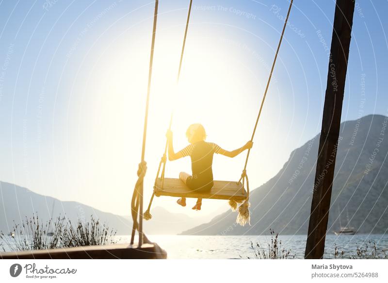 Little boy having fun on large swing on the shore in the Boka Kotor Bay of the Adriatic Sea in the Balkan Mountains, Montenegro. Happy child on a sea resort on sunny summer holidays day.