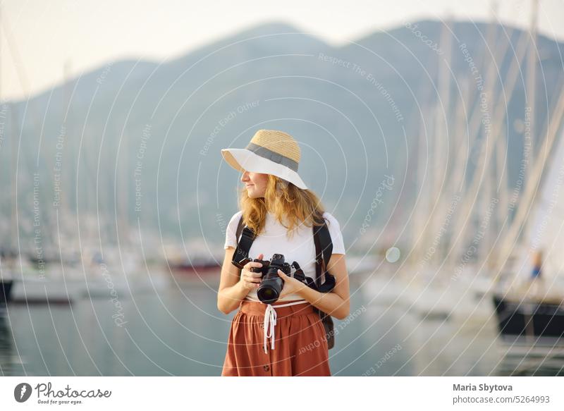 Charming young tourist woman taking a picture on the Mediterranean coast. Attractive red-haired girl photographer with a camera on the background of sea and yachts. Tourism and travel