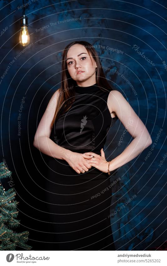 young beautiful woman in a black festive long dress near the Christmas tree. Elegant lady in black dress over christmas tree lights background. happy new year