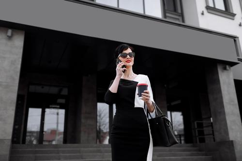 Successful businesswoman leader stands in big city talks on mobile phone. Smiling woman making business call on cell walking with cup of coffee on background of office centre, multitasking outdoors