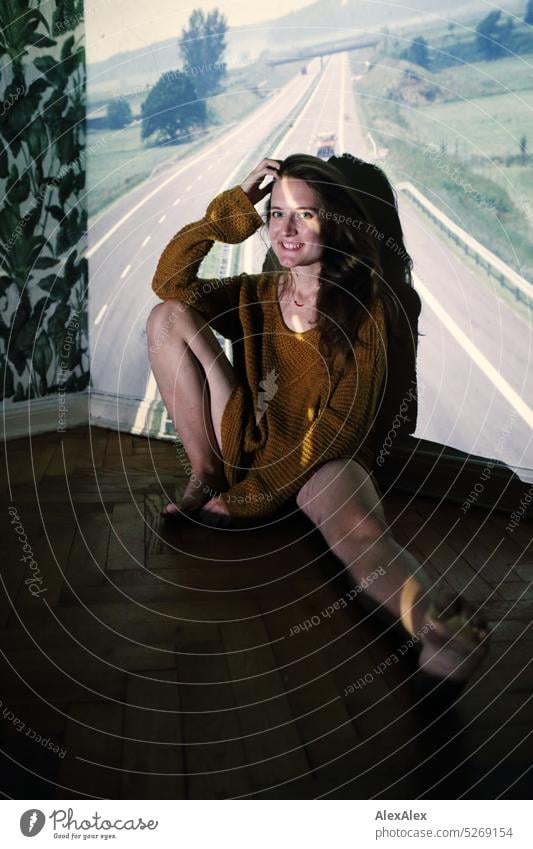Young woman sits with sweater and bare legs on parquet floor and is illuminated by an old slide showing a highway Woman Smiling Sweater Long-haired Slim