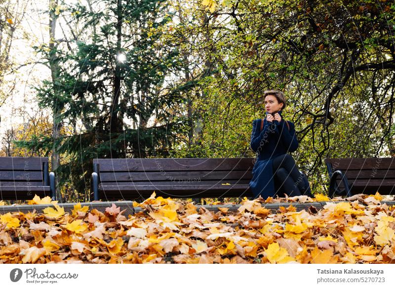 A woman wraps herself in a coat sitting on a bench in an autumn park fall female adult short hair Blond Contemplation Watching Anticipation Awaiting Attractive