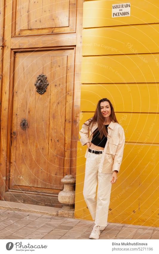 Young woman standing near yellow wall and wooden brown door looking in the camera millennial urban model fashion city tourist tourism young adult shy angry