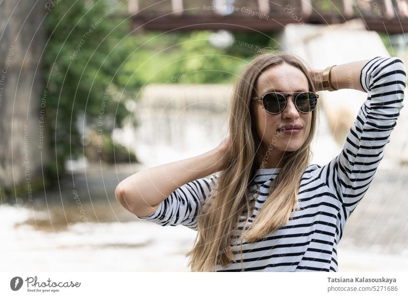 A woman in sunglasses and a striped top holds her hair near the river with her hands hands. longsleeve jumper dam female people person adult Mid adult women