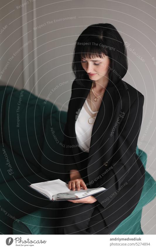 Beautiful business woman writing something in the note pad while sitting on the green sofa at office by window, charming brunette female with short hair in black suit working indoors