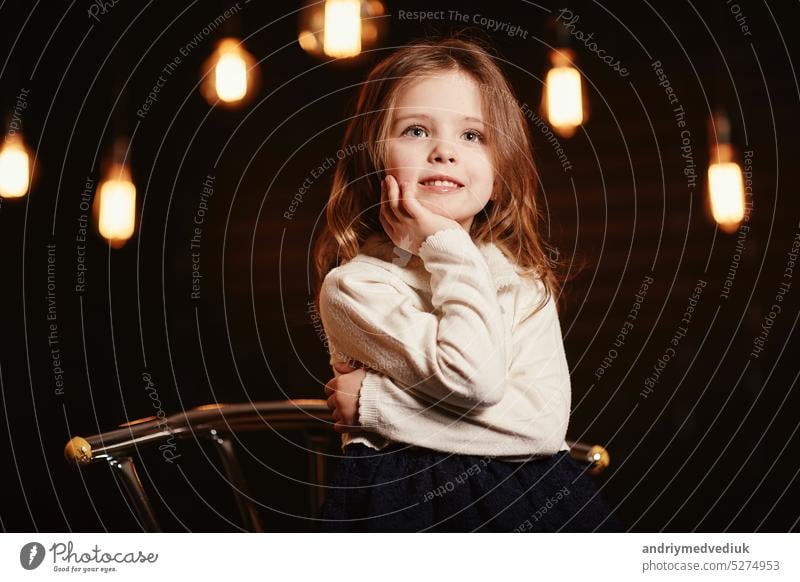 Little adorable smiling girl is touching face with her hands and looking in camera with a dark background. Child in lace blue dress and knit sweater is sitting on metal chair. Happy childhood