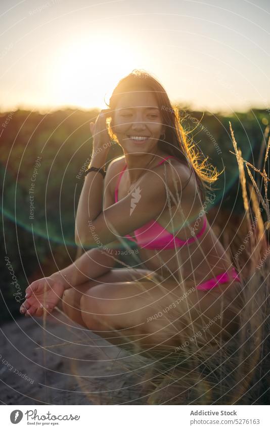 Cheerful female sitting on haunches on beach woman touch hair smile sunset summer happy weekend vacation resort young sundown cheerful coast glad swimsuit