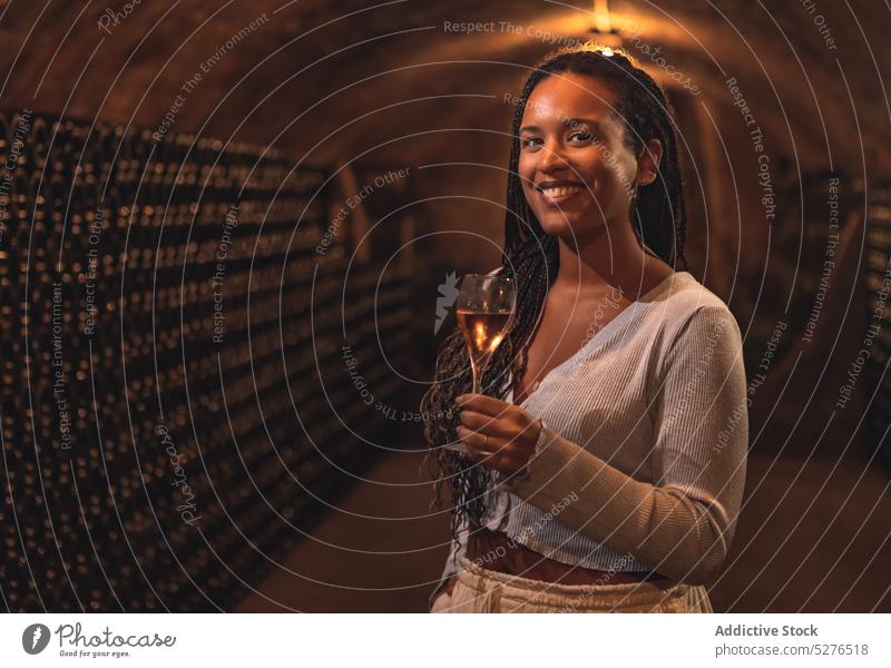 Joyful ethnic lady smiling while drinking wine in cellar woman smile alcohol confident winery enjoy portrait beverage bottle female young african american black
