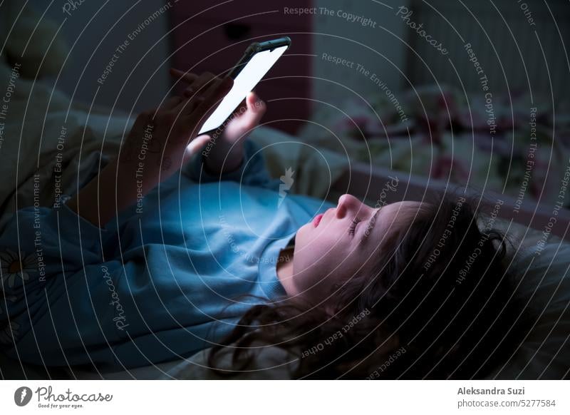 A child using smart phone lying in bed late at night, playing games, watching videos online, scrolling screen. Children's screen addiction. Child's room at night.