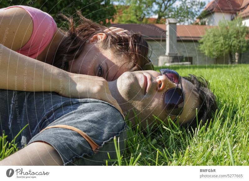 Upper body couple what is lying in the grass in summer with T shirt. She is cuddled up to him and looks into the camera while he wears sunglasses and lies with his head in the grass.