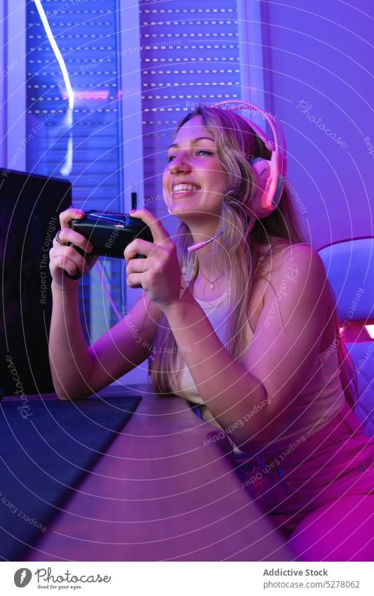 Cheerful female streamer in headphones using mic and gamepad woman cheerful console play happy microphone record blogger neon room headset talk young illuminate