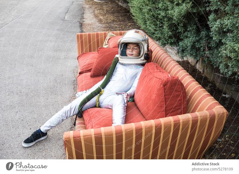 Child in space suit relaxing on sofa on street boy child spaceman cosmonaut astronaut spacesuit sleep couch chill rest kid explorer helmet costume park preteen