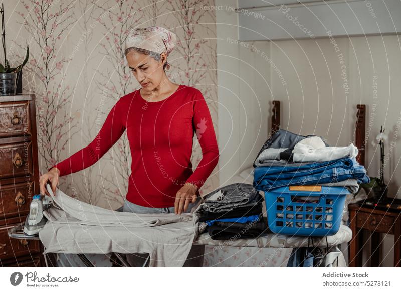 Mature woman folding clothes in room iron household home housework domestic routine chore ethnic housewife aged prepare female calm outfit senior focus shirt