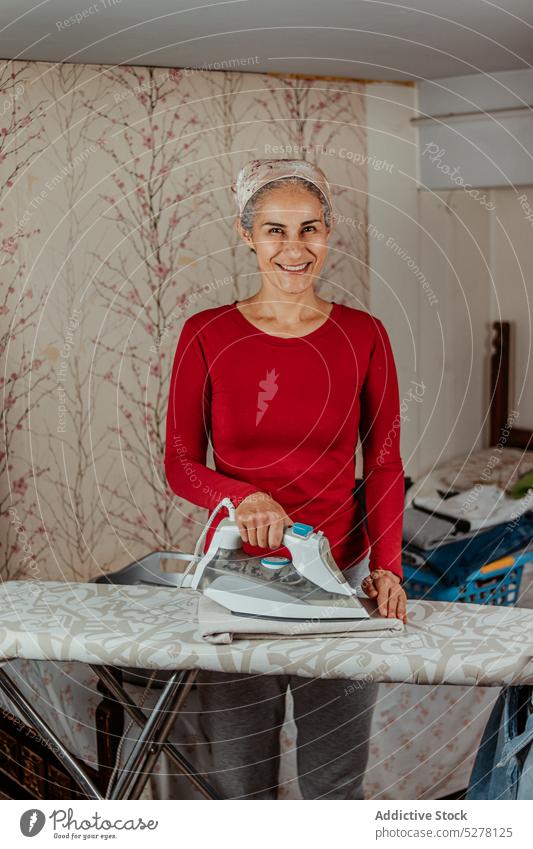 Smiling woman ironing clothes in room home household housework routine chore housewife female domestic daily apartment duty garment lifestyle appliance flat