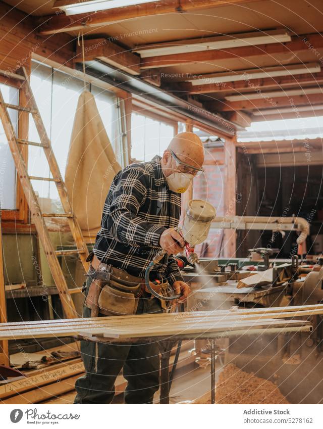 Carpenter using paint sprayer in workshop man varnish carpenter board woodworker mask joinery polish plank concentrate professional goggles male focus protect