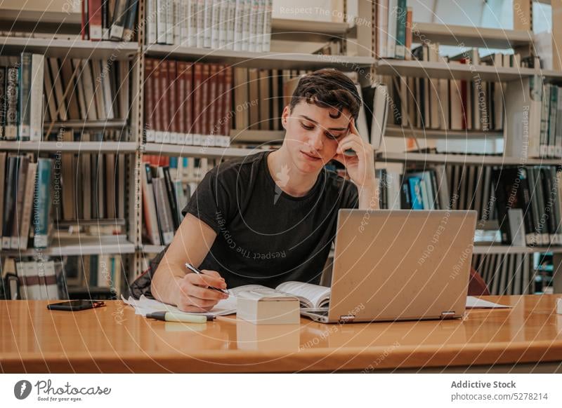 Focused male student preparing for exam in library man homework laptop write take note knowledge concentrate study education college learn exam preparation
