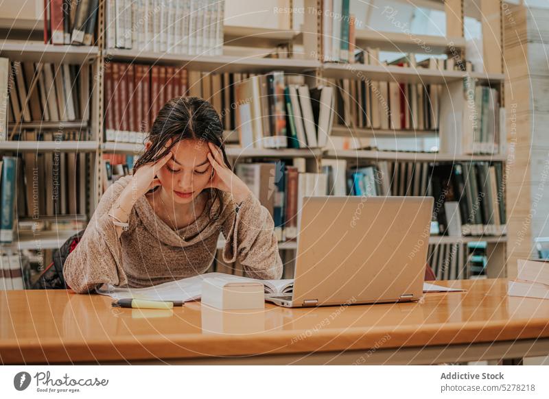 Pensive female student reading book in library woman study laptop education homework exam preparation learn knowledge research thoughtful young pensive