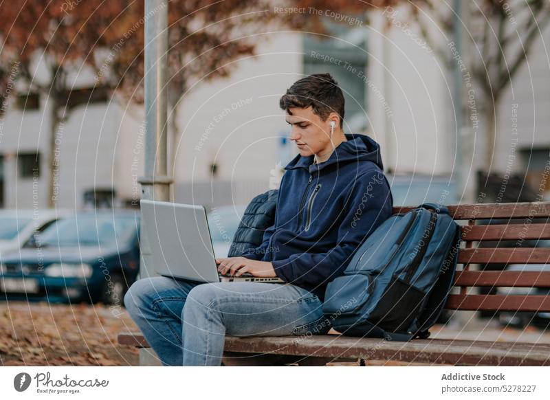 Concentrated man browsing laptop on bench earphones park student autumn focus concentrate fall young male warm clothes gadget device backpack internet surfing