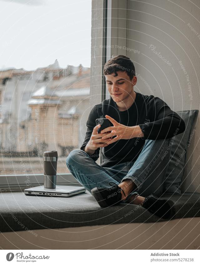Smiling teenager browsing smartphone on windowsill man student break tea laptop cheerful smile message male legs crossed positive device surfing chat casual