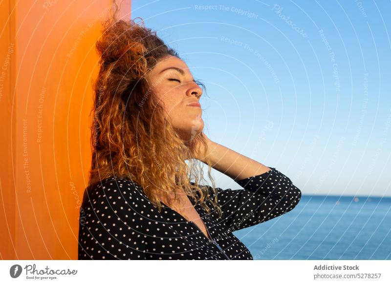 Pensive woman touching curly hair against sea touch hair freedom wind street summer sunlight leisure individuality calm tranquil female young appearance adjust