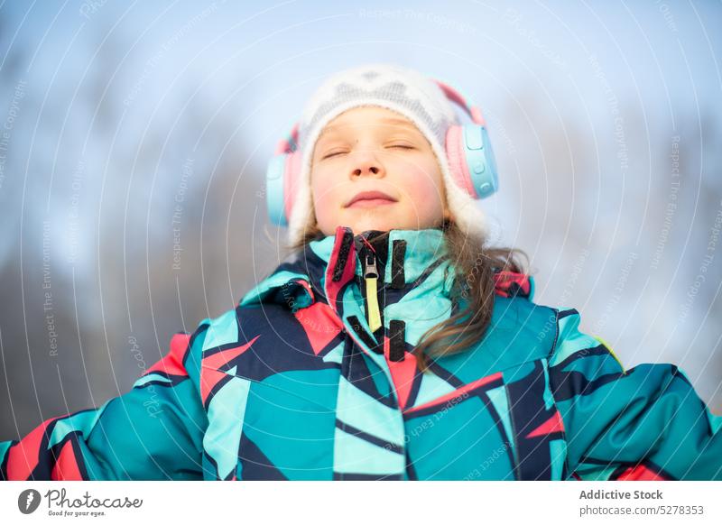 Calm girl listening to music in headphones winter content eyes closed warm clothes cold smile hat melody outerwear kid audio song enjoy season street sound