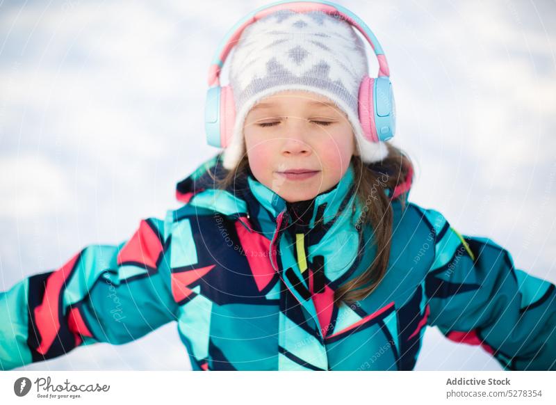 Calm girl listening to music in headphones winter content eyes closed warm clothes cold smile hat melody outerwear kid audio song enjoy season street sound