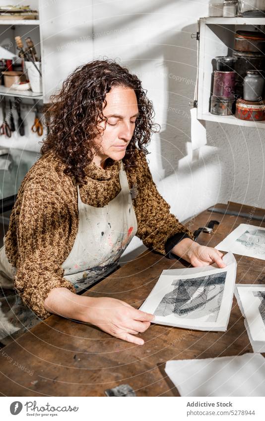 Female artisan inspecting engraved photos craftswoman examine photoengraving paper handmade table studio female middle age mature workshop hobby handicraft