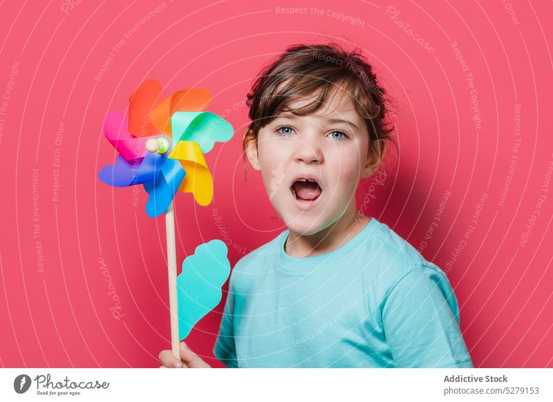Cute girl holding pinwheel and looking at camera kid rainbow flower petal play open mouth leisure entertain funny playful toy child individuality joy