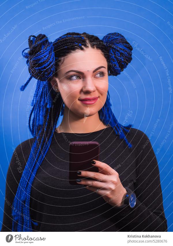 Adorable hipster caucasian female with african colorful braids hairstyle standing with mobile phone, smiling, posing on blue wall beautiful girl portrait pretty