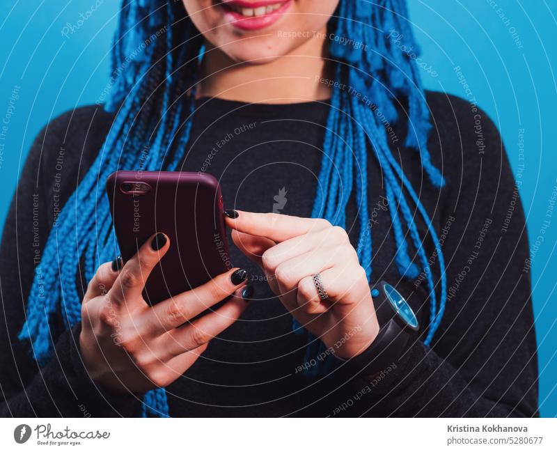 Adorable hipster caucasian female with african colorful braids hairstyle checking news feed or messaging via social networks, using free wi-fi on mobile phone, smiling, posing on blue wall