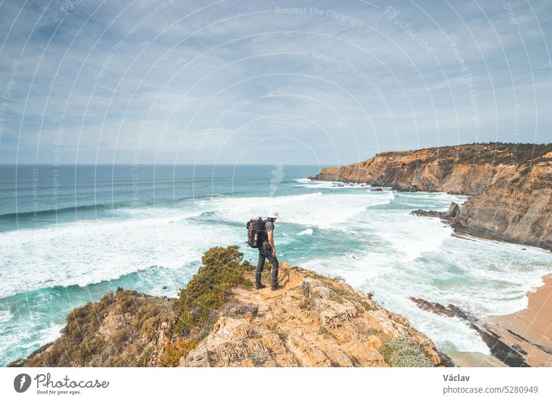 Hiker and adventurer stand on a rugged rock and cliff coastline and enjoy the view on the Atlantic Ocean in the west of Portugal in the famous tourist region of the Algarve