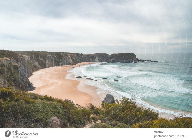 Famous Praia dos Machados beach in Odemira, western Portugal. The rocks surrounding the moon-shaped sandy beach with large waves from the Atlantic Ocean. Fisherman Trail